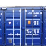A blue shipping container with identification numbers "CPIU 034082 2261" and various caution labels on the doors. The self storage container has multiple locks securing the doors and is situated on a gravel surface.