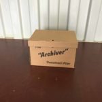 A closed brown cardboard box labeled "The 'Archiver' Document Filer" sits on a brown surface against a white corrugated metal wall background, perfect for personal storage or student storage needs.
