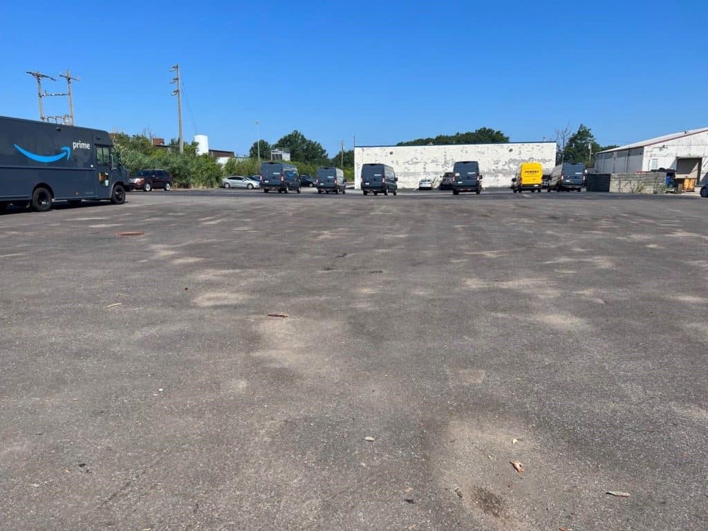 A large, mostly empty paved lot with a few delivery vans, including several Amazon Prime trucks, parked in a row near the back. Industrial buildings and a white wall with fading paint are visible in the background under a clear blue sky—ideal for business storage needs.