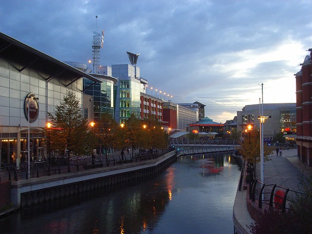 An urban canal scene at dusk features modern buildings with a mix of glass and brick architecture on either side of the water. Street lamps and building lights reflect on the canal water, while trees with autumn foliage line pedestrian paths, alongside strategically placed self storage containers for local businesses.