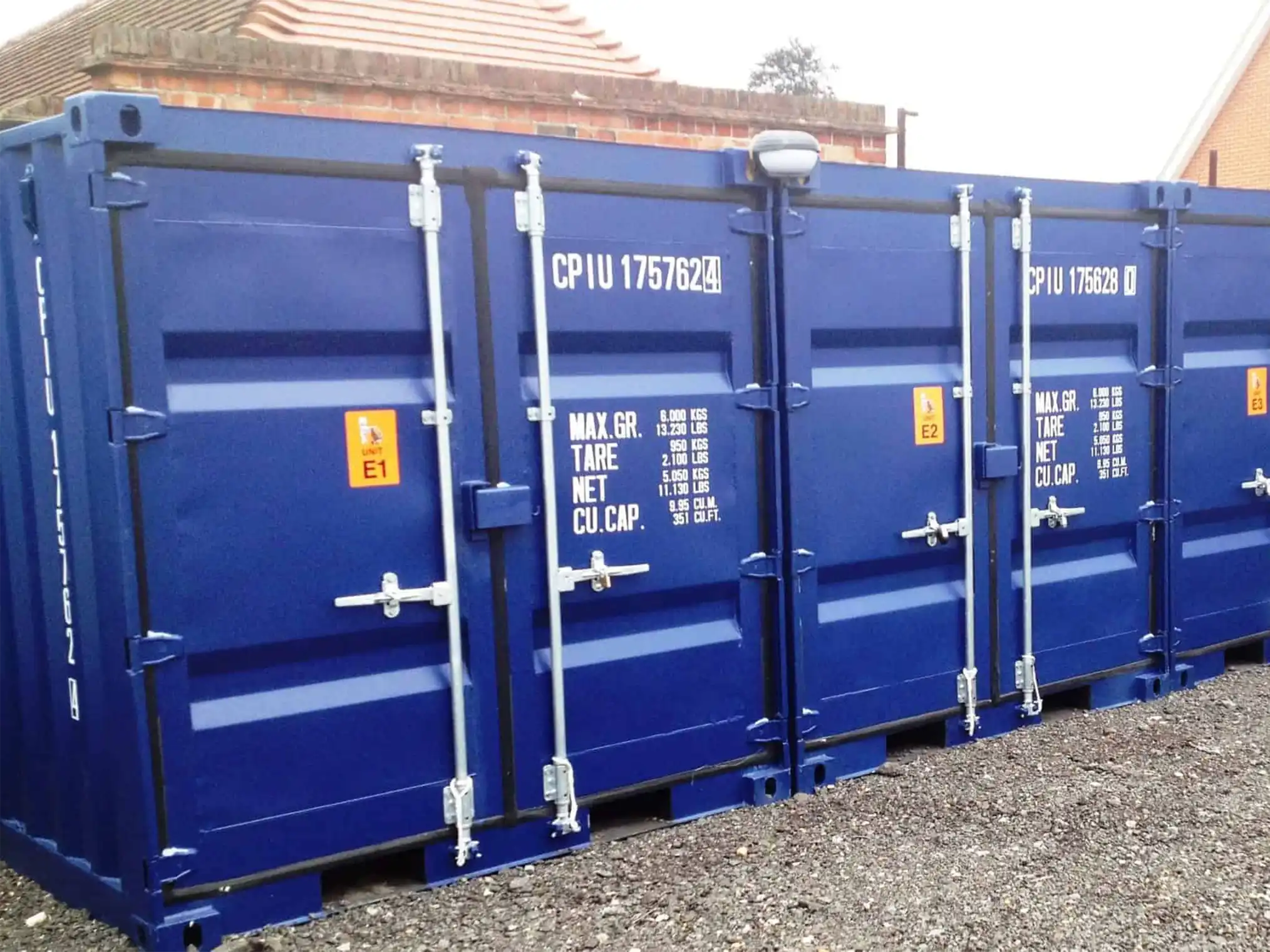 A row of blue storage containers with visible markings and serial numbers stands on a gravel surface. These self storage containers are secured with steel locking mechanisms and positioned near a building with brown brick walls.
