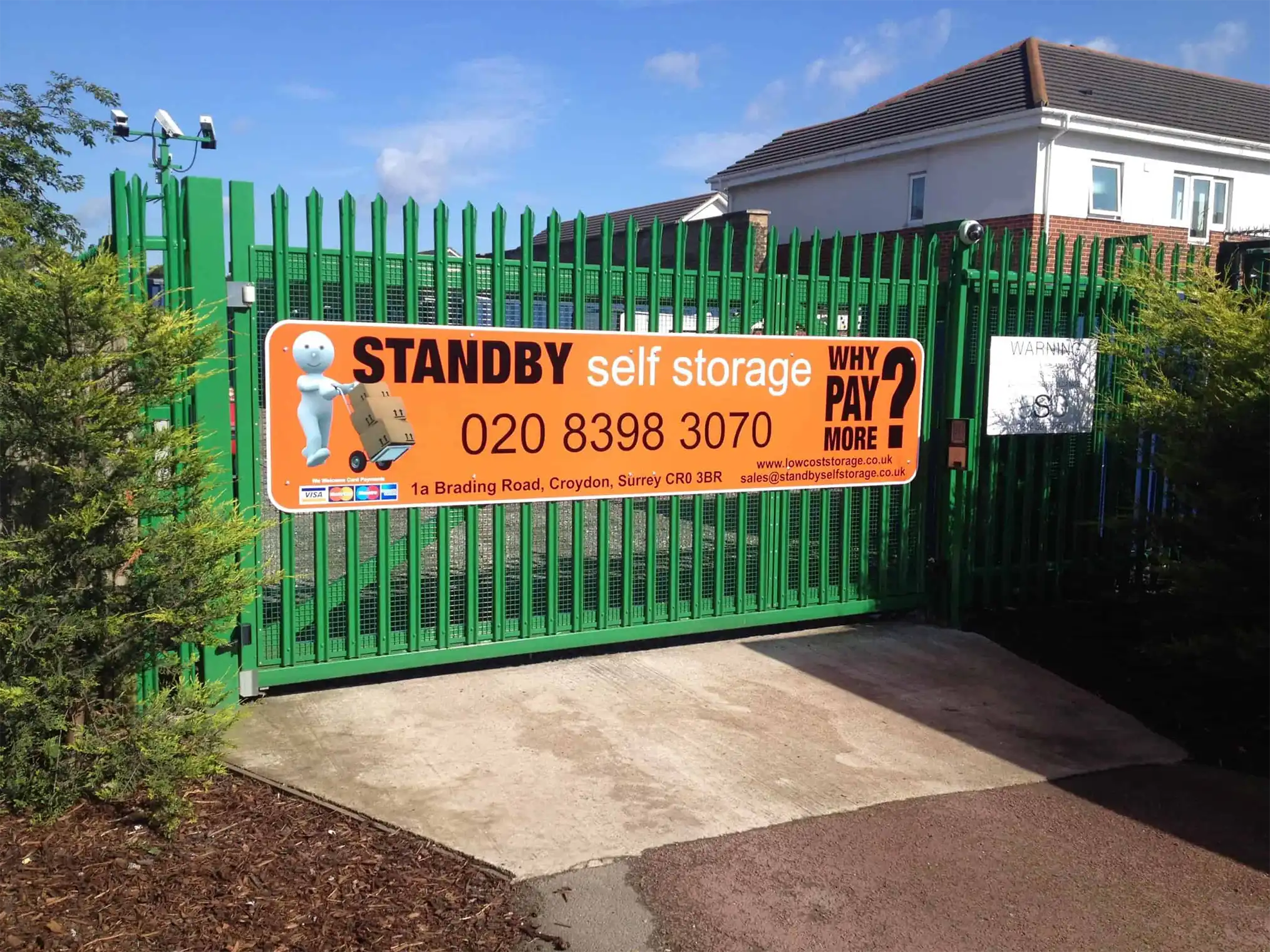 A green metal gate with a large orange sign promoting "Standby Self Storage." The sign includes contact information, a cartoon mascot, and the text "Why Pay More?" Perfect for student storage needs. A white house and a tree are visible in the background.