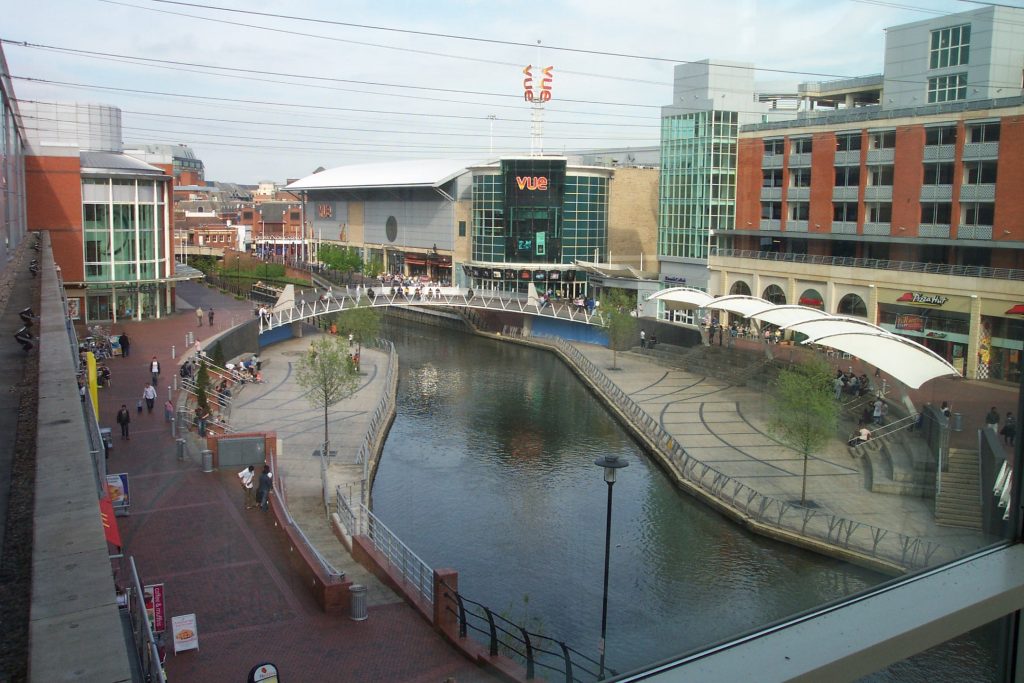 A canal flanked by modern buildings and pedestrian pathways in an urban shopping area. People walk on the pathways and sit near the water. A VUE cinema is visible, along with several shops and restaurants. Overhead tram lines stretch across the scene, blending harmoniously with neatly arranged student storage containers nearby.